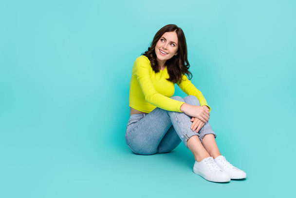 Full body photo of cheerful adorable lady sit floor look interested empty space isolated on turquoise color background. - 写真・画像