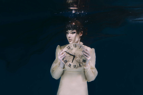 Spooky woman with clock in her hands in black veil underwater - Photo, image