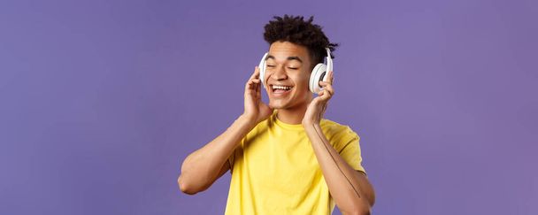 Close-up portret van zorgeloze, jonge vrolijke man met dreads, ogen dicht en glimlachend blij als het luisteren naar geweldige nieuwe lied favoriete artiest geüploade muziek platform, draag een koptelefoon. - Foto, afbeelding