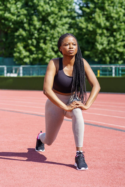 African American lady with long braids tries hard to pump gluteal muscles on red track of city stadium against lush green trees. Persistent woman takes care of fit body near park on summer day - Valokuva, kuva