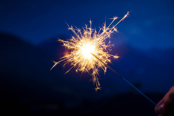Hand holding a sparkler, or bengal fire stick, burning in outdoor setting, mountain landscape, at dusk. Holidays or magic background or wallpaper. - 写真・画像