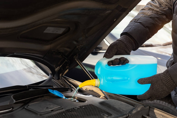 Close-up detail of driver or mechanic hand in gloves pouring blue antifreeze liquid for winter car windshield screen washing with bootle watering can. Car seasonal service and maintenance concept. - Photo, image
