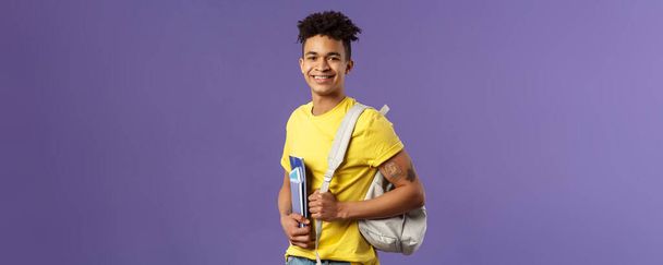 Back to school, university concept. Portrait of handsome charismatic hispanic guy, student walking to campus, going for lecture, studying hold backpack and notebooks, stand purple background. - Photo, image
