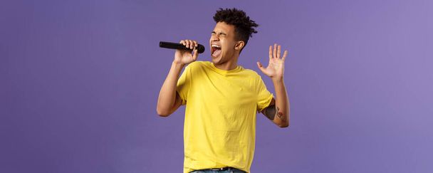 Retrato de un apasionado cantante hispano joven y despreocupado con temores y tatuajes, llegando a la nota más alta en la canción, levantando la mano cantando fuerte en el micrófono con los ojos cerrados, fondo púrpura
. - Foto, imagen