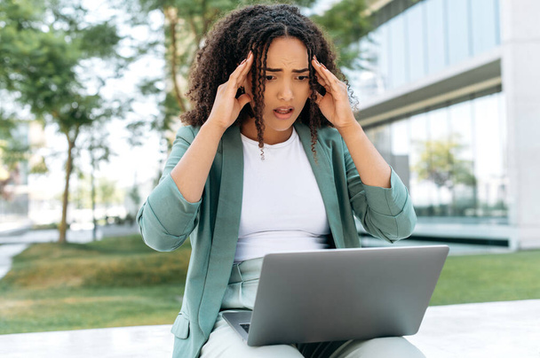 Disappointment puzzled brazilian or hispanic young business woman, elegantly dressed, sit outdoors with laptop, looking at screen in confusion, received unexpected news, holds her hands on her head - Foto, Imagem
