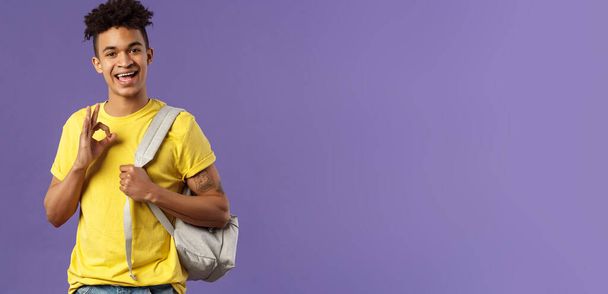 De vuelta a la escuela, concepto universitario. Retrato de joven chico hipster guapo, estudiante con mochila asegurar prueba ser bueno, mostrar signo de bien, relajarse y relajarse sin preocupaciones, todo bien, fondo púrpura
. - Foto, imagen