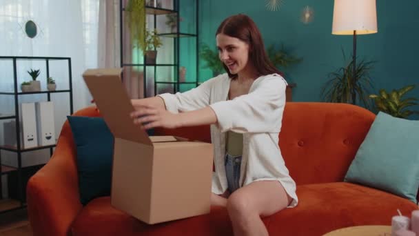 Πορτρέτο του ευτυχισμένου κοριτσιού ξεπακετάρισμα παράδοσης δεμάτων κάθεται στο σπίτι. Χαμογελώντας ικανοποιημένη νεαρή γυναίκα αγοραστής, online κατάστημα πελάτη άνοιγμα χαρτονένιο κουτί παραλαβή δώρου αγοράς από γρήγορη ταχυδρομική αποστολή - Πλάνα, βίντεο