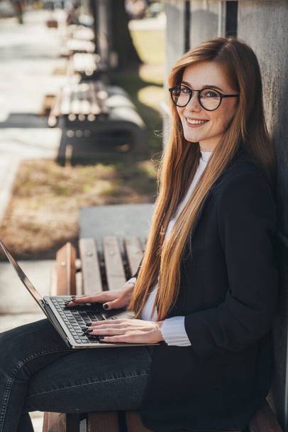 Young ginger student woman preparing for exams with laptop while sitting on wooden bench in the city park. Modern lifestyle. Summer vibes. Outdoor workplace. - Foto, Imagem