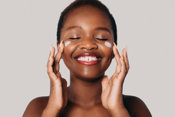 Front view portrait of an afro woman applying face cream onto her cheeks, smiling at camera with closed eyes, isolated over gray background. Healthy lifestyle - Fotó, kép