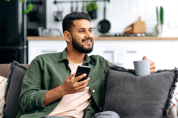 Happy indian or arabian attractive guy using his smartphone, sit on sofa in living room, browsing internet and social networks, texting with friends or family, reads news, holds a cup of coffee, smile - Foto, immagini