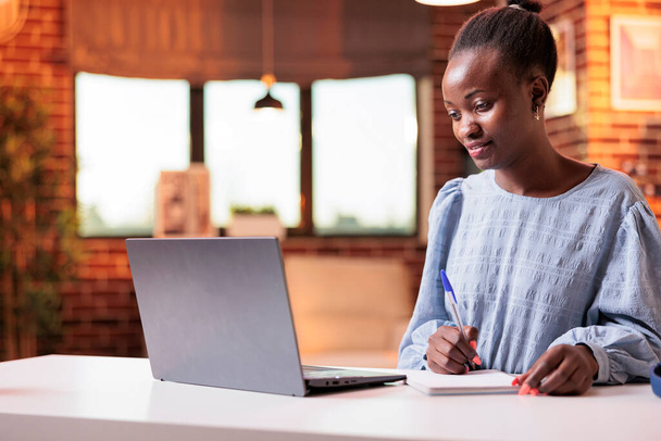 Woman studying with laptop and taking notes in notebook. Female african american remote university student watching educational tutorial and learning with online courses on internet - Foto, immagini