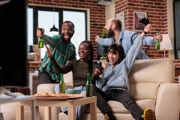 Happy friends playing video games and celebrating victory with beer bottles and pizza at fun gathering. Enjoying hangout at house party with group of people, leisure activity and win. - 写真・画像