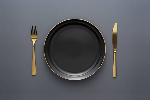 Золотые ножи и вилки на черном фоне, пустая черная тарелка. Красивые золотые столовые приборы. Вид сверху. - Фото, изображение