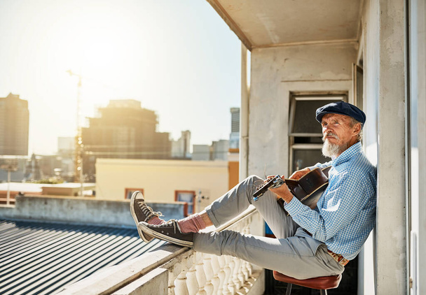 Φτιάχνει το τελευταίο του τραγούδι. ένας γαλήνιος ηλικιωμένος που κάθεται στο μπαλκόνι του παίζοντας ακουστική κιθάρα με την πόλη στο παρασκήνιο - Φωτογραφία, εικόνα