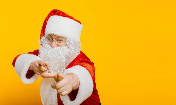 Santa claus points with index fingers at the camera on a yellow isolated background with copy space. - 写真・画像