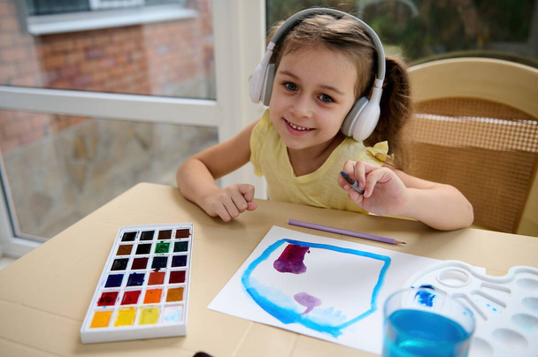 Adorable charming little girl of preschool wearing wireless headphones, smiles cutely looking at the camera, holding a paintbrush in her hand and enjoying watercolor art master class. Education, hobby - Fotografie, Obrázek