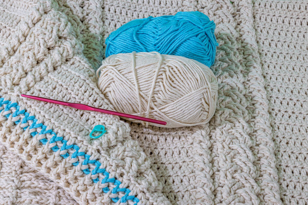 One beige and one blue skeins, pink metal hook and green stitch marker on a beige cotton yarn crochet blanket, intertwined braids, original raised crochet stitch pattern. Handmade craft creativity - Zdjęcie, obraz