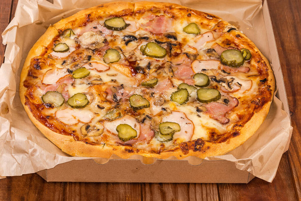 Round pizza with different ham, mushrooms and pickled cucumbers served in open cardboard pizza box on the old rustic table - Zdjęcie, obraz