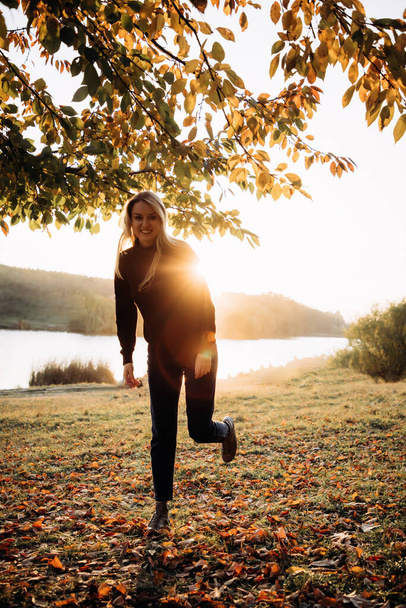 Beautiful girl with long blond hair in autumn landscape at sunset. Autumn portrait of a girl with the sun behind her, front view - Foto, Imagen