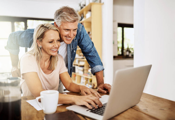 Счастливая, улыбающаяся и зрелая пара, использующая ноутбук вместе дома. Очаровательный муж помогает жене с интернет-работой. Веселые партнеры среднего возраста работают в команде в социальных сетях - Фото, изображение