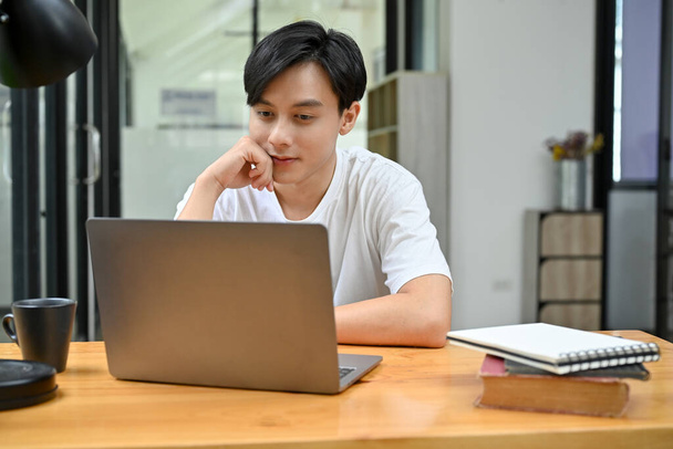 Relaxed and casual young Asian male college student or male office employee working in the office, using laptop computer, looking something on screen, thinking and planning his project. - Photo, Image