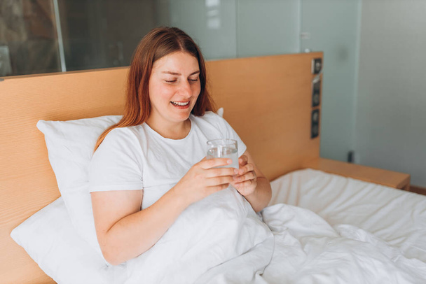 Healthy beautiful young woman holding glass of water on bed at home. Hand holds glass of clear water. Concept of healthy life - Photo, Image