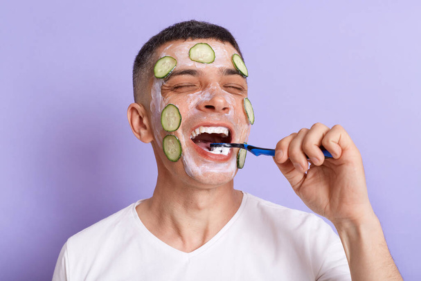 Positive happy man wearing white t shirt applying mask with cucumber slices on his face brushing his teeth, holding toothbrush in hands, keeps eyes closed, isolated over purple background. - Photo, image