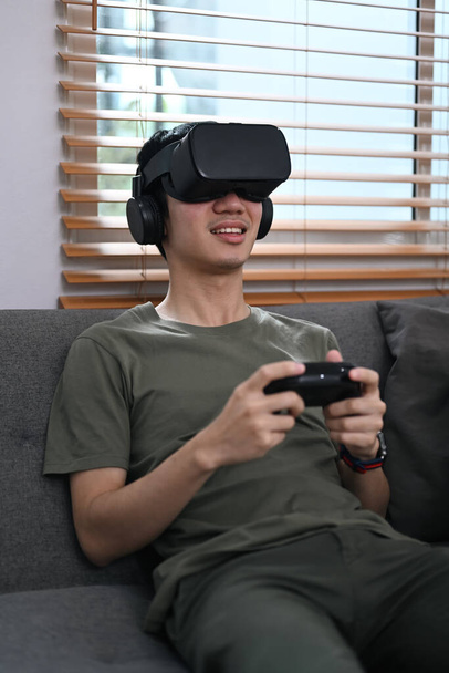 Ενθουσιασμένος άνθρωπος κρατώντας χειριστήριο και παίζοντας βιντεοπαιχνίδι με γυαλιά εικονικής πραγματικότητας στον καναπέ. - Φωτογραφία, εικόνα