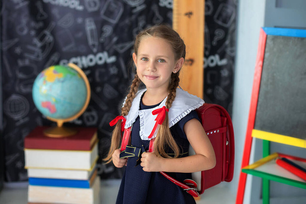Girl Child with red backpack studies in classroom against background of black board in school. Smiling Little girl with pigtails and in a school uniform stands classroom at school. Back to school  - Foto, immagini