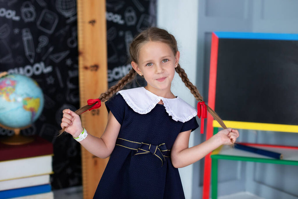 Smiling Little girl with pigtails in her hands and in a school uniform stands classroom at school. Child plays and studies in classroom against background of black board in school. Back to school  - Fotó, kép