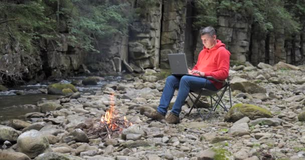 A man works on a laptop near a stream in nature. Concept of freelancing, digital nomad or remote office. 4K - Metraje, vídeo