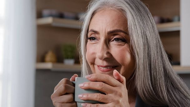 Dreamy inspired old blonde grey-haired woman granny grandmother mature middle aged woman pensioner sitting in kitchen drinking coffee tea hot drink healthy liquid from cup daydreaming relaxing at home - Photo, image