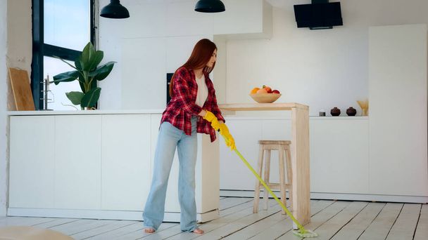 Young neat housewife redhead woman with long hair girl from cleanup agency take care of home house cleanliness washes floor with mop cleaning apartment modern kitchen room wipes wet scrubbing parquet - Fotoğraf, Görsel