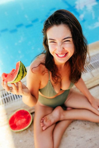 Cheerful young woman holding slice of watermelon in her hand, sitting near the pool and looking at camera with wide smile. Summer tastes. - Fotografie, Obrázek