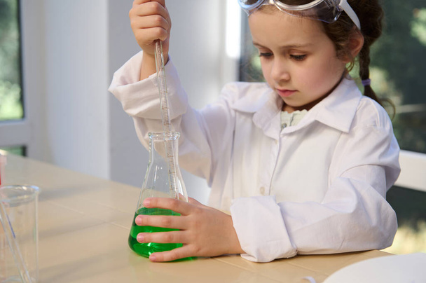 Adorable preschooler girl wearing white lab coat, enjoying learns chemistry at home laboratory, using flask and pipette, makes experiment . Fascinating scientific experiments for kids entertainment - Photo, image