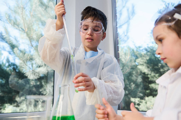 Красивый кавказский подросток, умный школьник в лабораторной защитной одежде, используя пипетку, закапывает реагент в пробирку, стоит рядом со своим одноклассником, наблюдая за происходящей химической реакцией - Фото, изображение