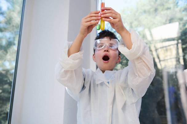 Focus on the hands of a schoolboy in white lab coat and glasses holding laboratory test tubes and observing the chemical reaction taking place in them, while performing experiments at chemistry lesson - Fotoğraf, Görsel