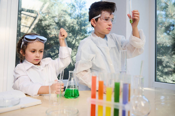 Diverse kids, teenage boy and preschooler girl in white laboratory coats and protective eyeglasses, doing biochemistry research in chemistry class. Test tubes, flasks with chemicals on the foreground - Foto, immagini