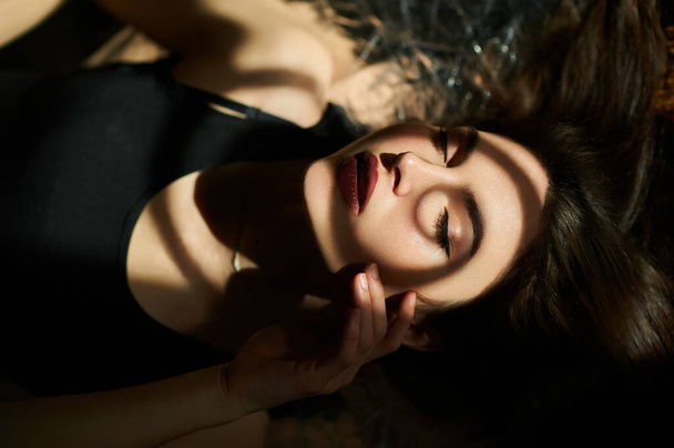 Portrait of a young girl lying with her eyes closed, with a shadow pattern on her face, touching the skin with her beautiful cheek. - Foto, Bild