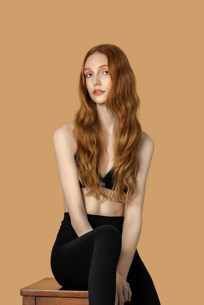 Portrait of a beautiful young White woman with long red hair, light makeup and lipstick sitting by herself on top of a table inside a studio with a pecan background wearing a black bra top and leggings. - Foto, afbeelding