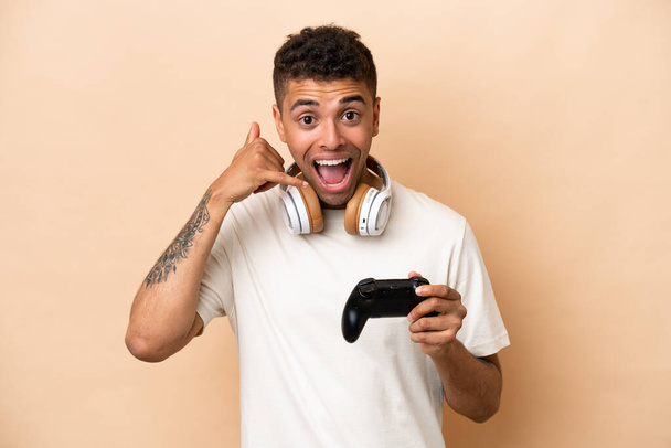 Młody Brazylijczyk bawiący się kontrolerem gier wideo na beżowym tle, wykonujący gest telefoniczny. Oddzwoń. - Zdjęcie, obraz