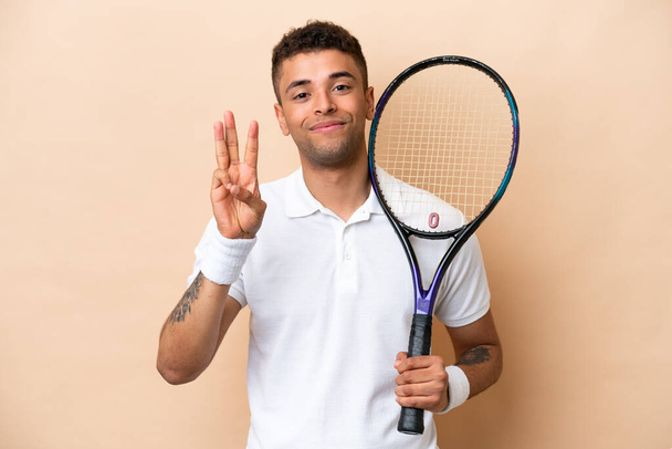 Jovem brasileiro bonitão jogando tênis isolado em fundo bege feliz e contando três com os dedos - Foto, Imagem