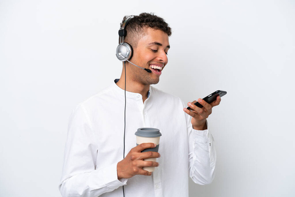 Telemarketer brasilianische Mann arbeitet mit einem Headset isoliert auf weißem Hintergrund hält Kaffee zum Mitnehmen und ein Handy - Foto, Bild