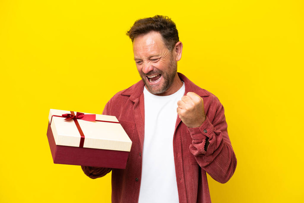 Middle age caucasian man holding a gift isolated on yellow background celebrating a victory - Photo, Image