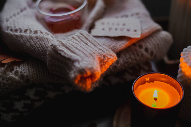 Two burning candles, glass cup of tea and stack of warm cozy sweaters om bedtable. Cozy autumn evening at home. Selective focus - Foto, afbeelding