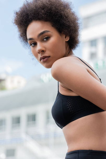 athletic african american woman in black sports bra looking at camera outdoors - Zdjęcie, obraz