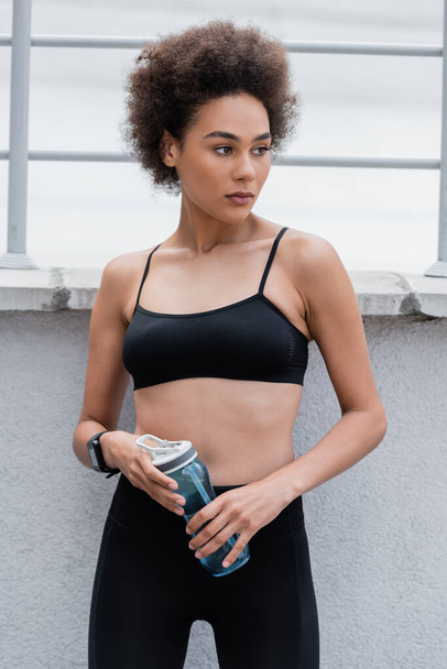 pretty african american woman in black sports bra holding sports bottle and looking away - Valokuva, kuva