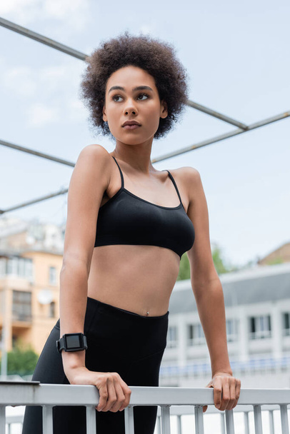 curly african american woman in black sports bra and fitness tracker looking away near fence - Fotografie, Obrázek