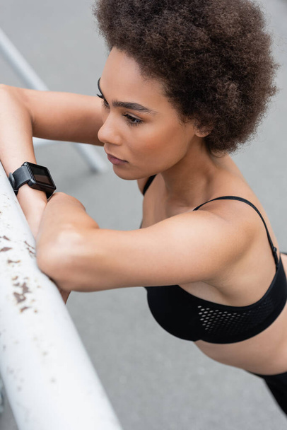 high angle view of curly african american woman in fitness tracker leaning on fence outdoors - Fotografie, Obrázek