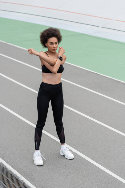 full length of african american woman in black sportswear and white sneakers warming up on stadium - Foto, afbeelding